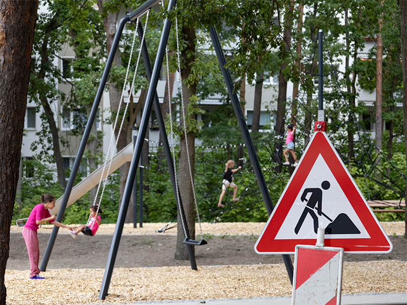 17.6.2026, Baustellenschild und Warnbarke, im Hintergrund spielen die ersten Kinder auf den neuen Spielanlagen